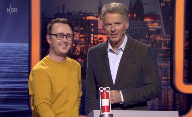 Zwei Männer stehen nebeneinander, einer trägt ein gelbes Sweatshirt. Hintergrund mit Stadtansicht.