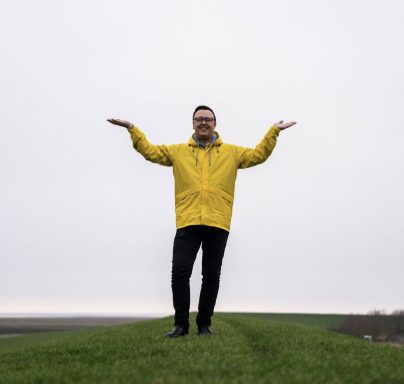 Mann in gelber Jacke steht auf einer Wiese, Hände ausgebreitet.