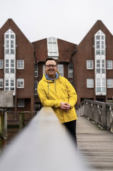 Mann in gelbem Regenmantel steht auf einem Holzsteg vor einem roten Backsteingebäude.