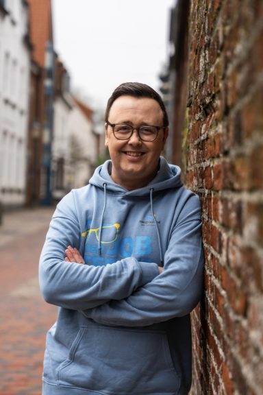 Mann in blauem Hoodie steht an einer Ziegelmauer in einer Stadtstraße.