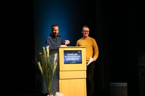 Zwei Männer stehen an einem Rednerpult während einer Präsentation auf der Bühne.