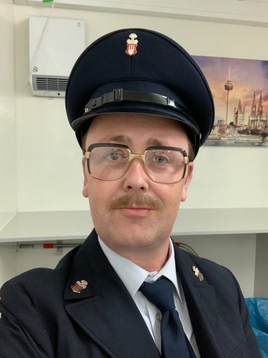 Ein Mann in Uniform mit einer Mütze und Brille, lächelt in die Kamera.
