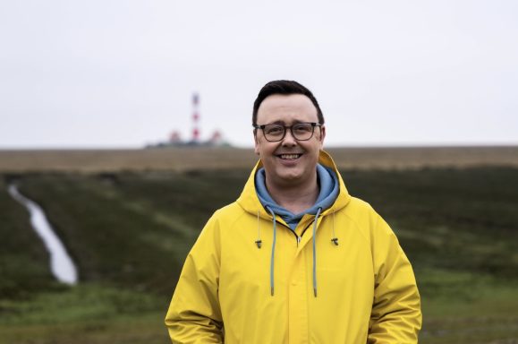 Mann in gelber Jacke steht lächelnd in einer Landschaft mit einem Industriebau im Hintergrund.
