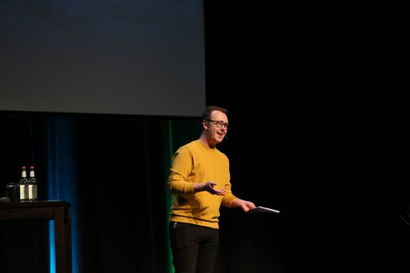 Person in einem gelben Pullover spricht auf einer Bühne, Hintergrund mit Bildschirm.